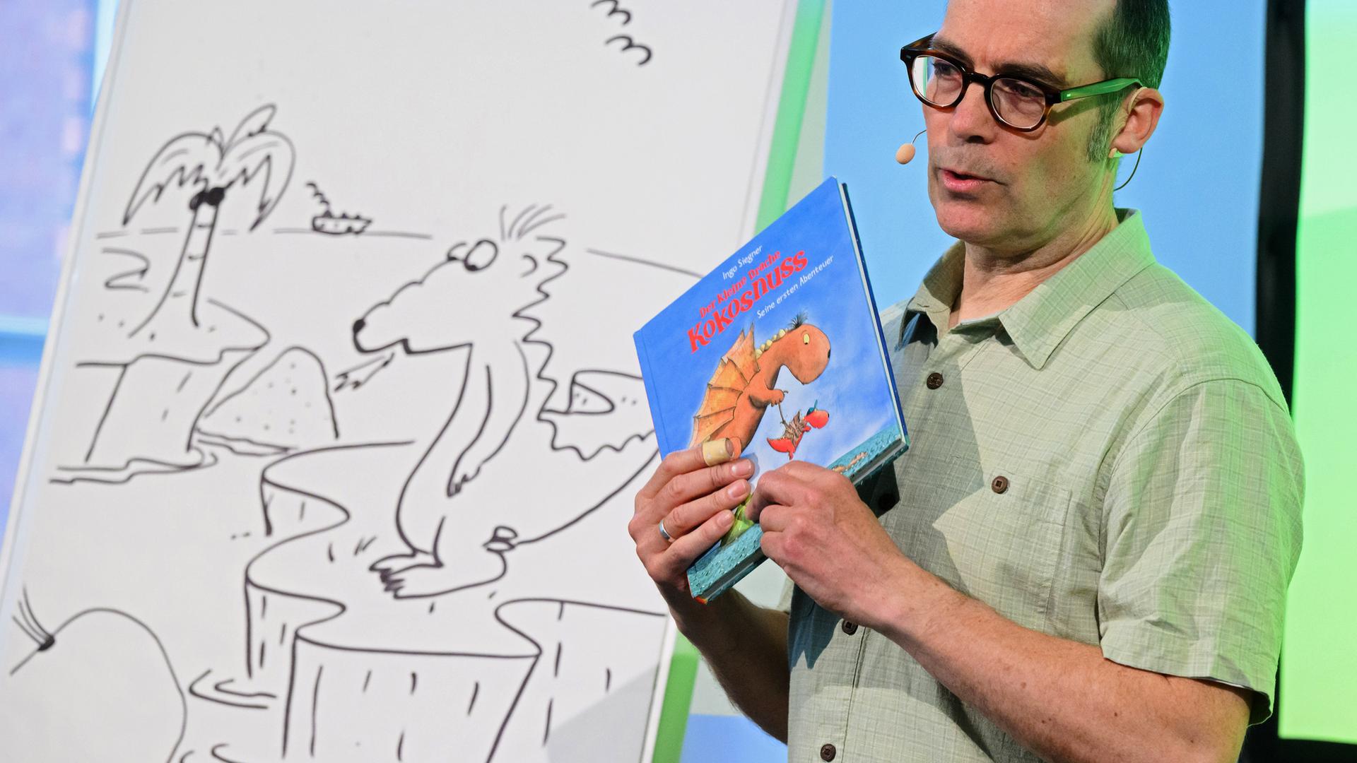 Links steht ein Whiteboard, auf das ein Drache, eine Palme und Vögel gemalt wurden. Daneben steht ein Mann, der ein Buch in der Hand hält, auf dem auch ein Drache zu sehen ist. 