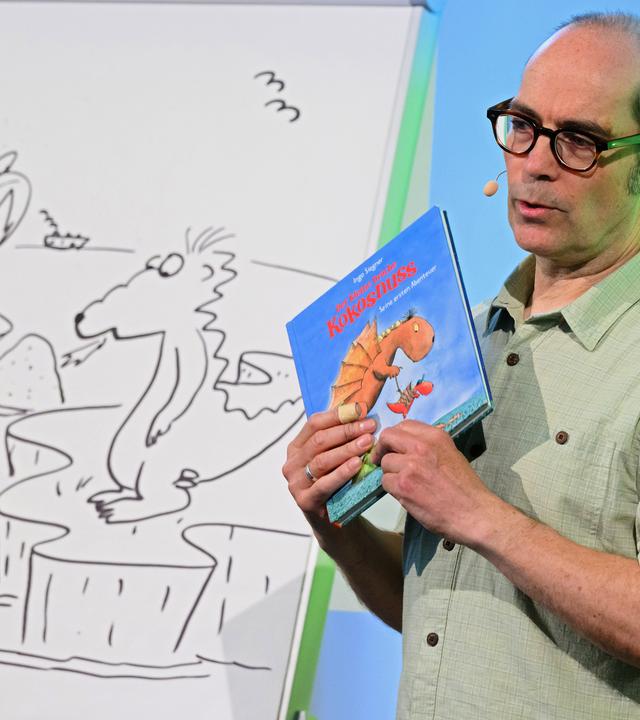 Links steht ein Whiteboard, auf das ein Drache, eine Palme und Vögel gemalt wurden. Daneben steht ein Mann, der ein Buch in der Hand hält, auf dem auch ein Drache zu sehen ist. 