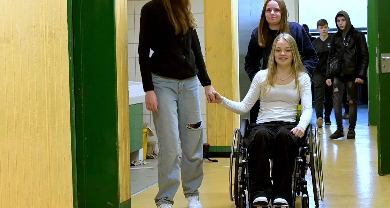 Mädchen im Rollstuhl wird von einer Mitschülerin geschoben, während eine andere Mitschülerin sie an der Hand hält