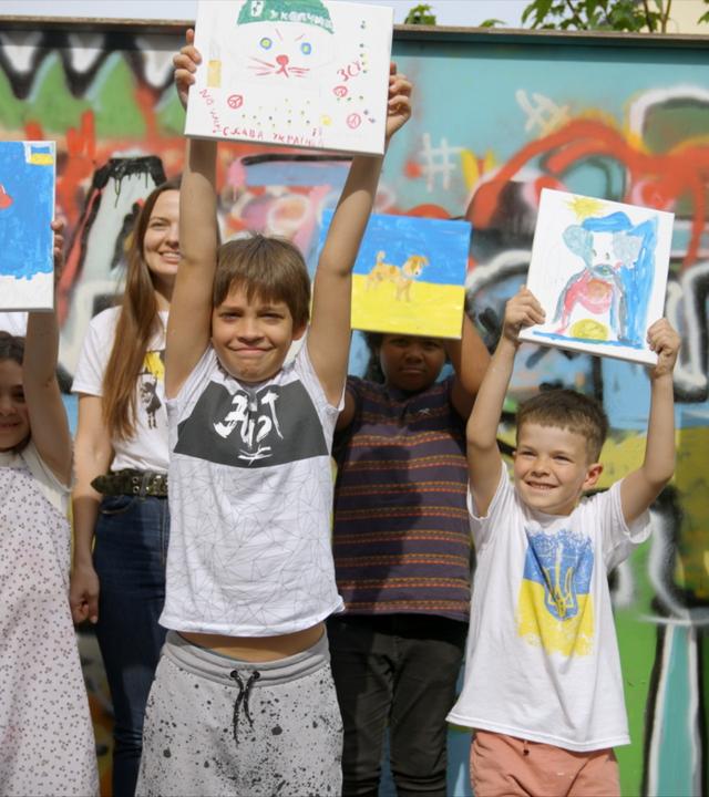 Eine Gruppe fröhlicher Kinder zeigt stolz ihre bunten Kunstwerke vor einer farbenfrohen Graffitiwand.