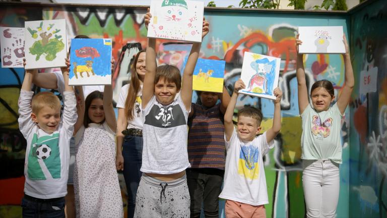 Eine Gruppe fröhlicher Kinder zeigt stolz ihre bunten Kunstwerke vor einer farbenfrohen Graffitiwand.