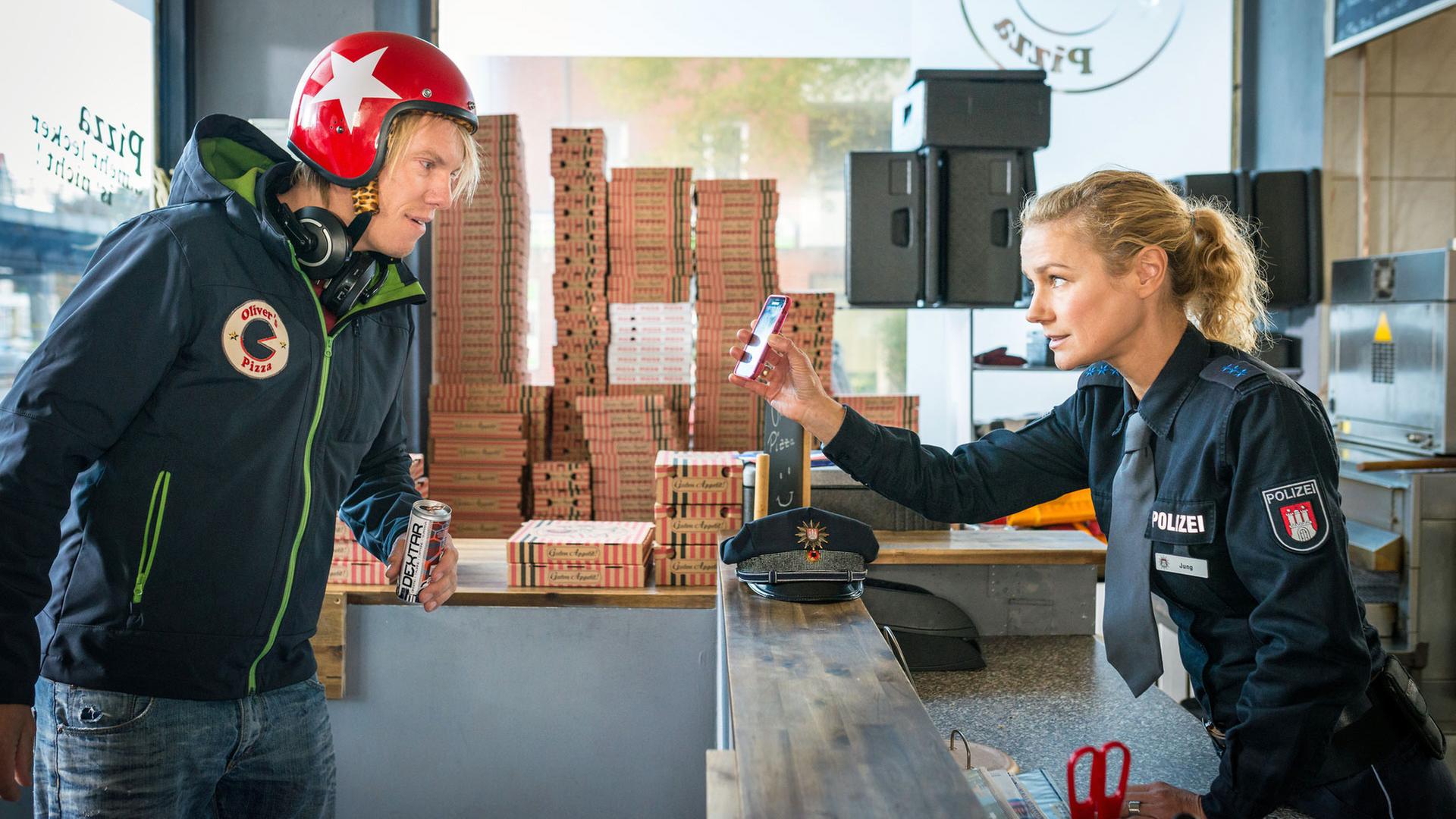 "Notruf Hafenkante - Innere Werte": Die Polizistin Franzi Jung (Rhea Harder-Vennewald) steht in einer Schnellpizza hinter der Theke und zeigt dem Pizzaboten (Simon Gosejohann), der vor der Theke steht ihr Handy. Sie schaut ihn dabei an, er schaut auf das Handy. Im Hintergrund stehen Pizzakartons gestapelt.