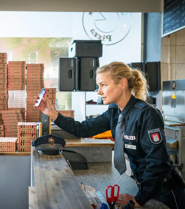 "Notruf Hafenkante - Innere Werte": Die Polizistin Franzi Jung (Rhea Harder-Vennewald) steht in einer Schnellpizza hinter der Theke und zeigt dem Pizzaboten (Simon Gosejohann), der vor der Theke steht ihr Handy. Sie schaut ihn dabei an, er schaut auf das Handy. Im Hintergrund stehen Pizzakartons gestapelt.