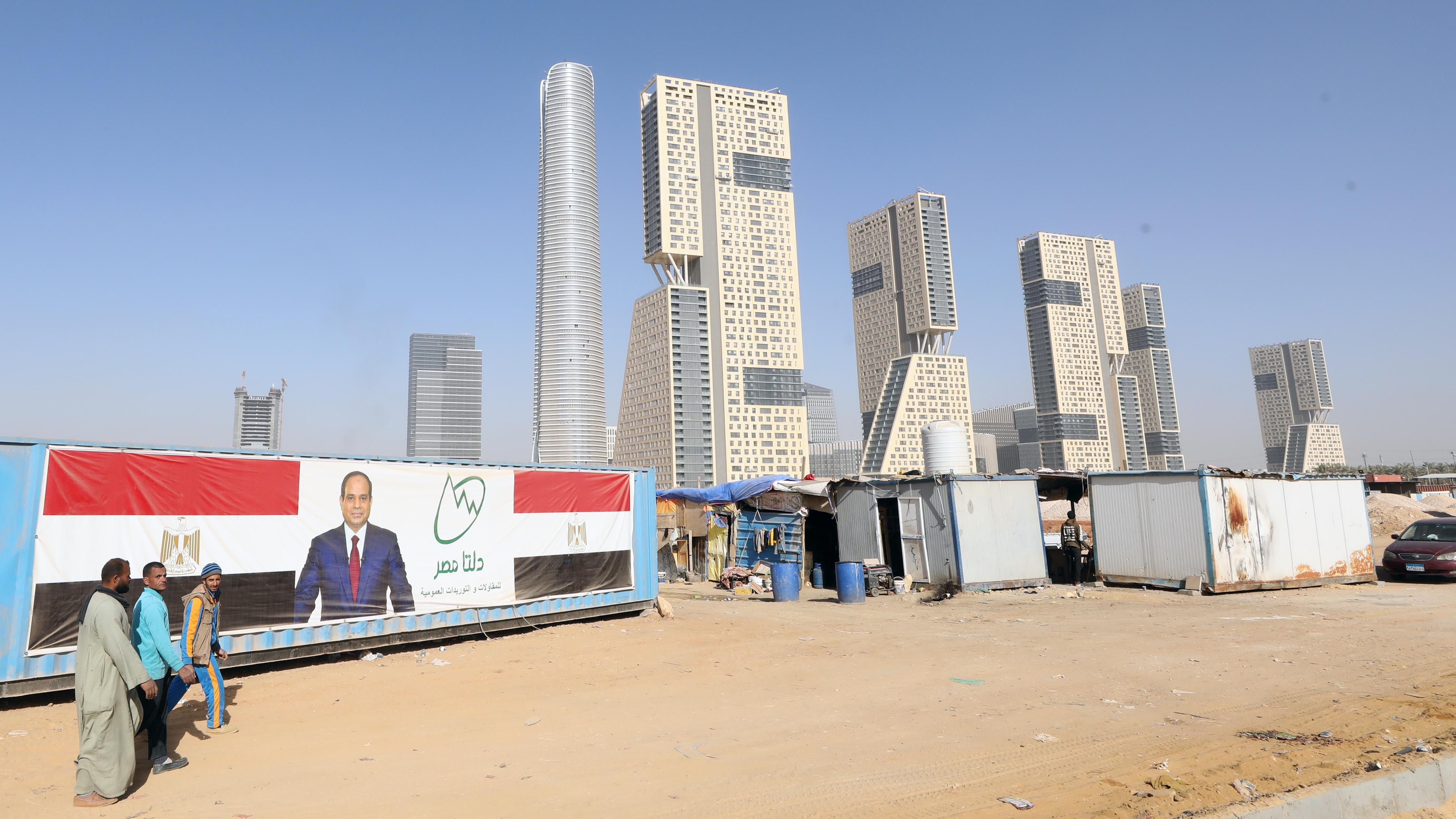 Neue Hochhäuser in der Wüste, Teil der neuen ägyptischen Hauptstadt.Davor stehen rechts Arbeiter baracken links ein großes Plakat mit ägyptischer Flagge und President Abdel Fattah al-Sisi.