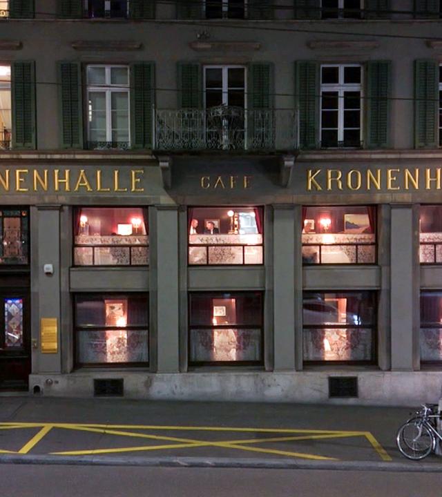 Außenansicht des traditionsreichen Restaurants Kronenhalle in Zürich bei Nacht, beleuchtete Fenster und Schriftzug gut sichtbar, davor ein geparktes Auto und zwei Fahrräder.