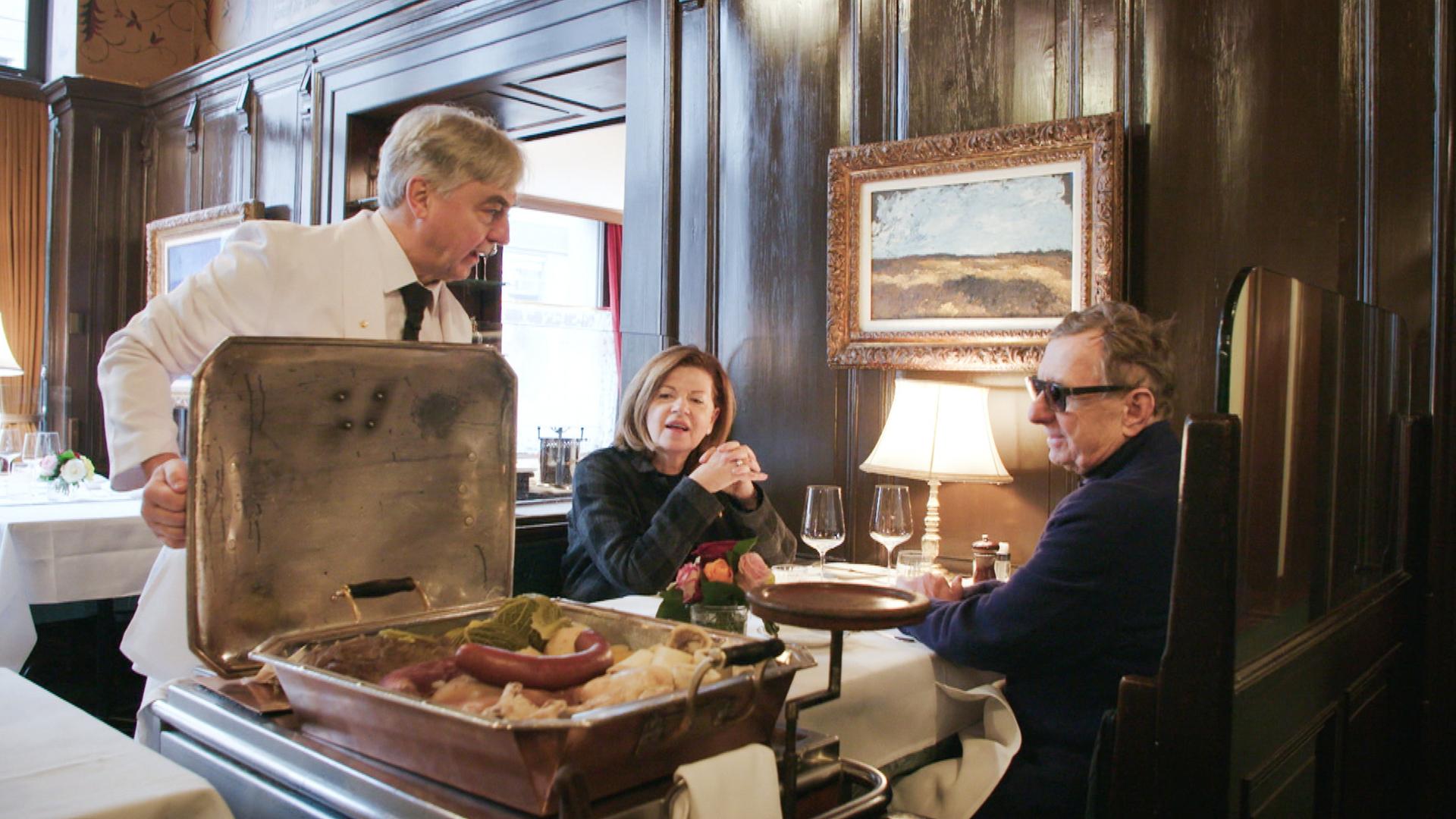 Ein Kellner mit weißem Jackett serviert einem Paar in der Kronenhalle Zürich traditionelle Speisen vom Speisewagen in edlem Ambiente mit dunkler Holzvertäfelung.