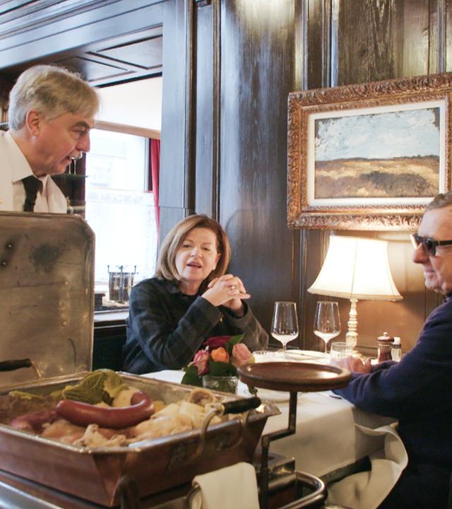 Ein Kellner mit weißem Jackett serviert einem Paar in der Kronenhalle Zürich traditionelle Speisen vom Speisewagen in edlem Ambiente mit dunkler Holzvertäfelung.
