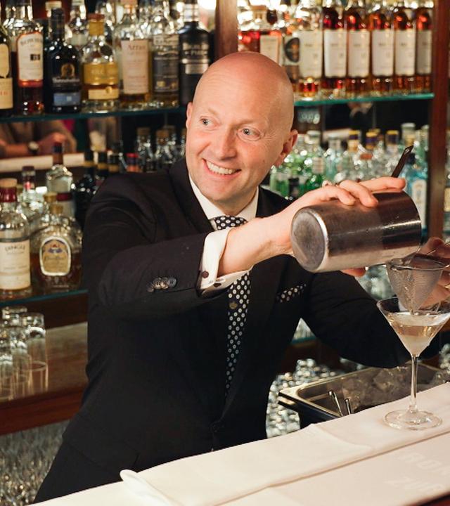 Christian Heiss, Chef de Bar in der Kronenhalle-Bar, mixt lächelnd einen Drink vor einer umfangreichen Sammlung edler Spirituosenflaschen.