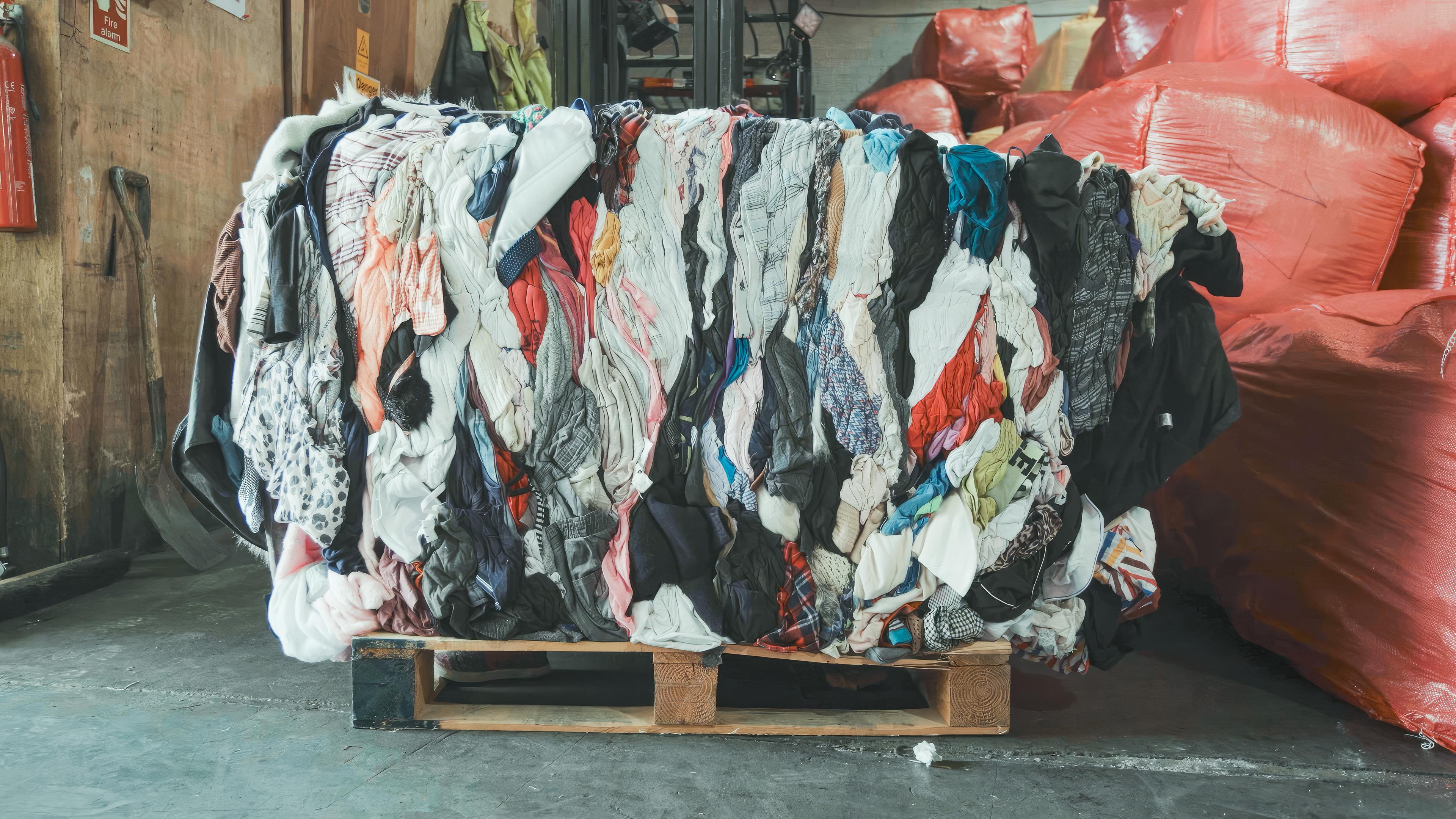 Ein großer Altkleider-Haufen steht zusammengepresst auf einer Europalette, daneben große, rote Säcke voller Altkleider.