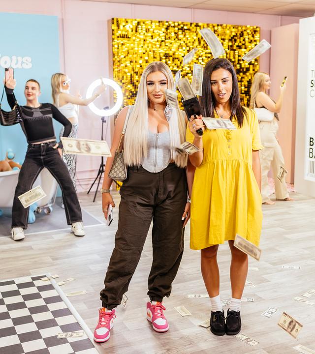 Zwei Frauen posieren vor der Kamera, eine hält ein Gerät in der Hand, aus dem Geldscheine fliegen. Im Hintergrund befinden sich fünf weitere Personen.