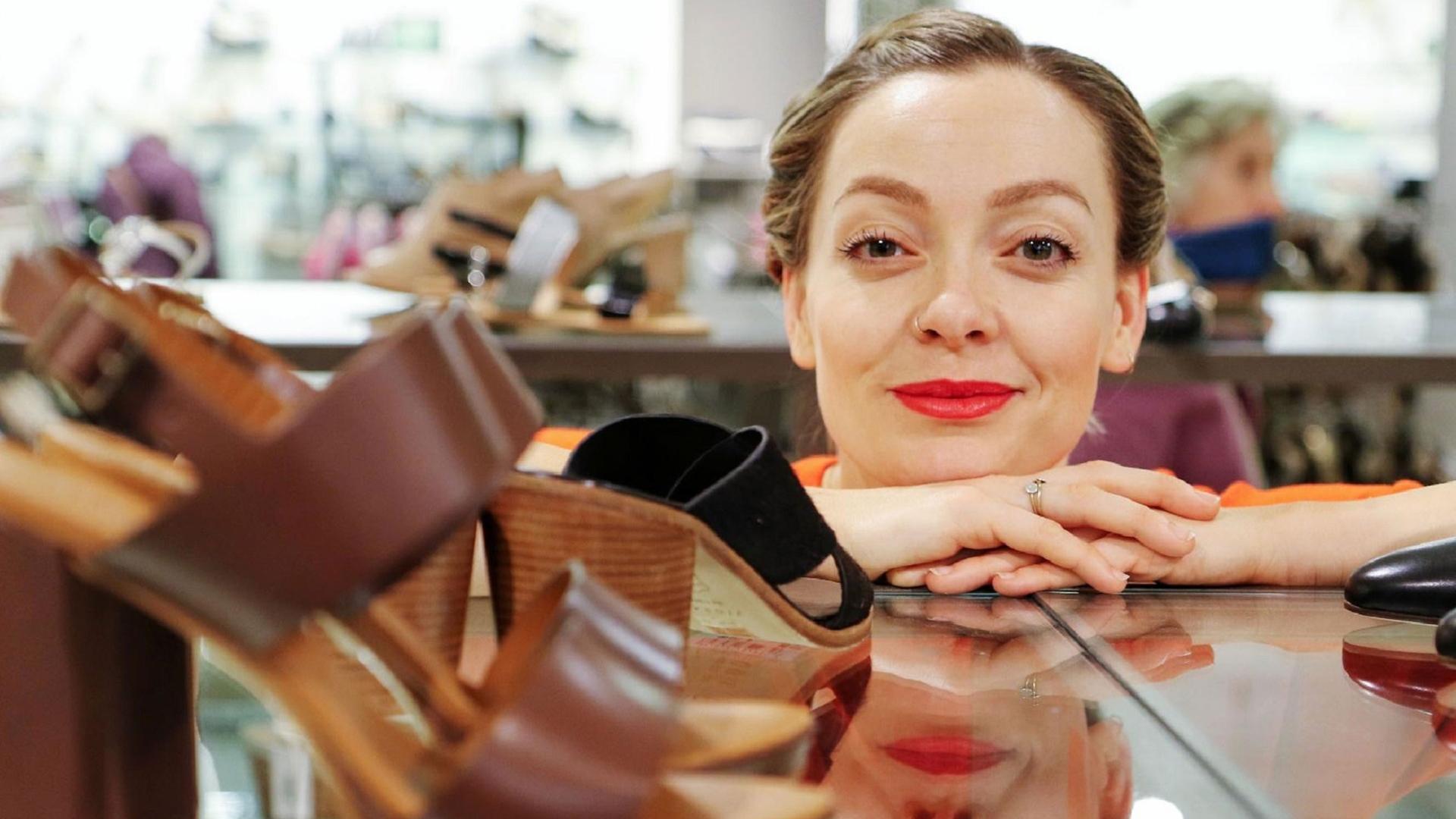 Cherry Healey steht hinter einem Schuhregal und schaut in die Kamera.