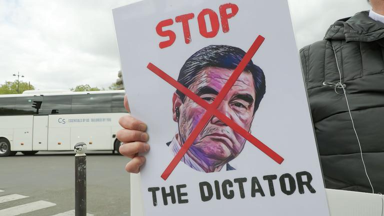 Auf der Straße vor einem Bus. Ein Arm hält ein Plakat mit durchgestrichenem Konterfei von Berdimuhamedow und der Aufschrift "Stop the Dictator".