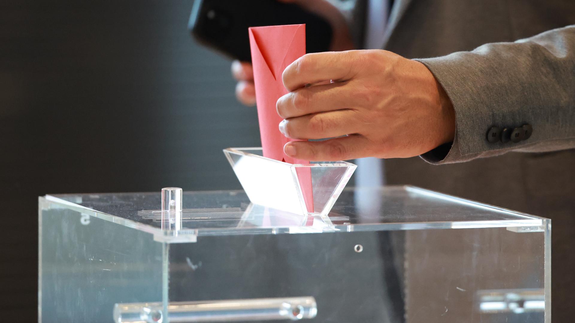 Typical für Wahlvorgänge: Eine Hand wirft einen Stimmzettel in eine transparente Wahlurne