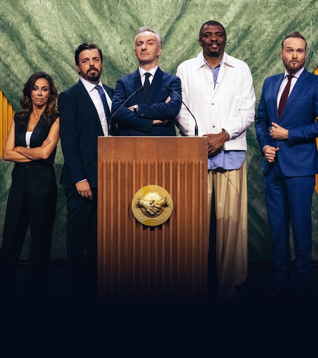 Von links nach rechts: Filomena Cautela, Anders Helenius, Jan Böhmermann, Loyiso Gola und Arjen Lubach stehen hinter einem großen Rednerpult. Im Hintergrund sind zahlreiche Landesflaggen in einer Reihe zu sehen.