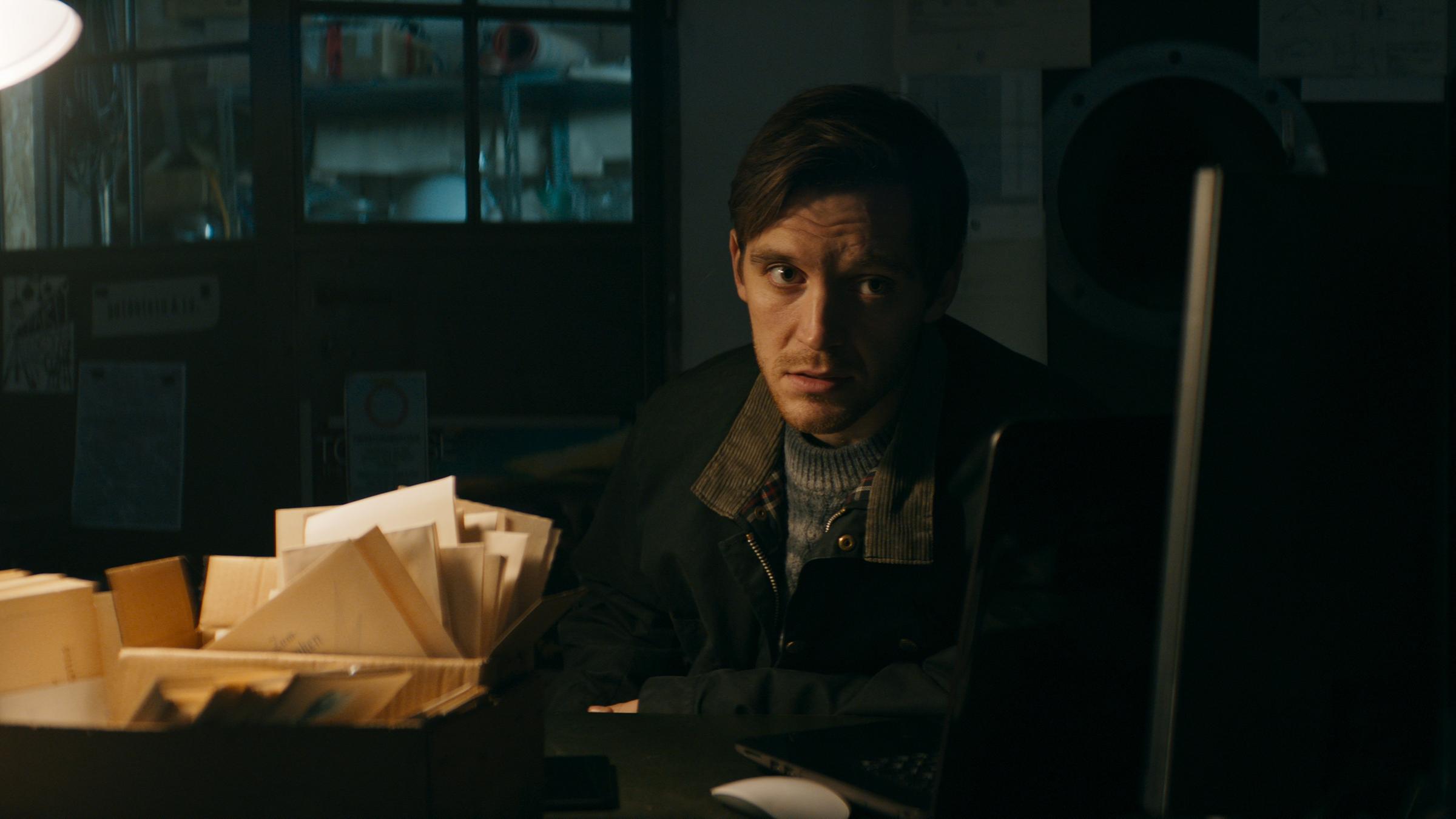 Bild aus der Serie Concordia: Leon (Jonas Nay), ein junger Mann in Jacke und Pulli, sitzt in einem dunklen Büro am Schreibtisch. Vor ihm Papiere. 