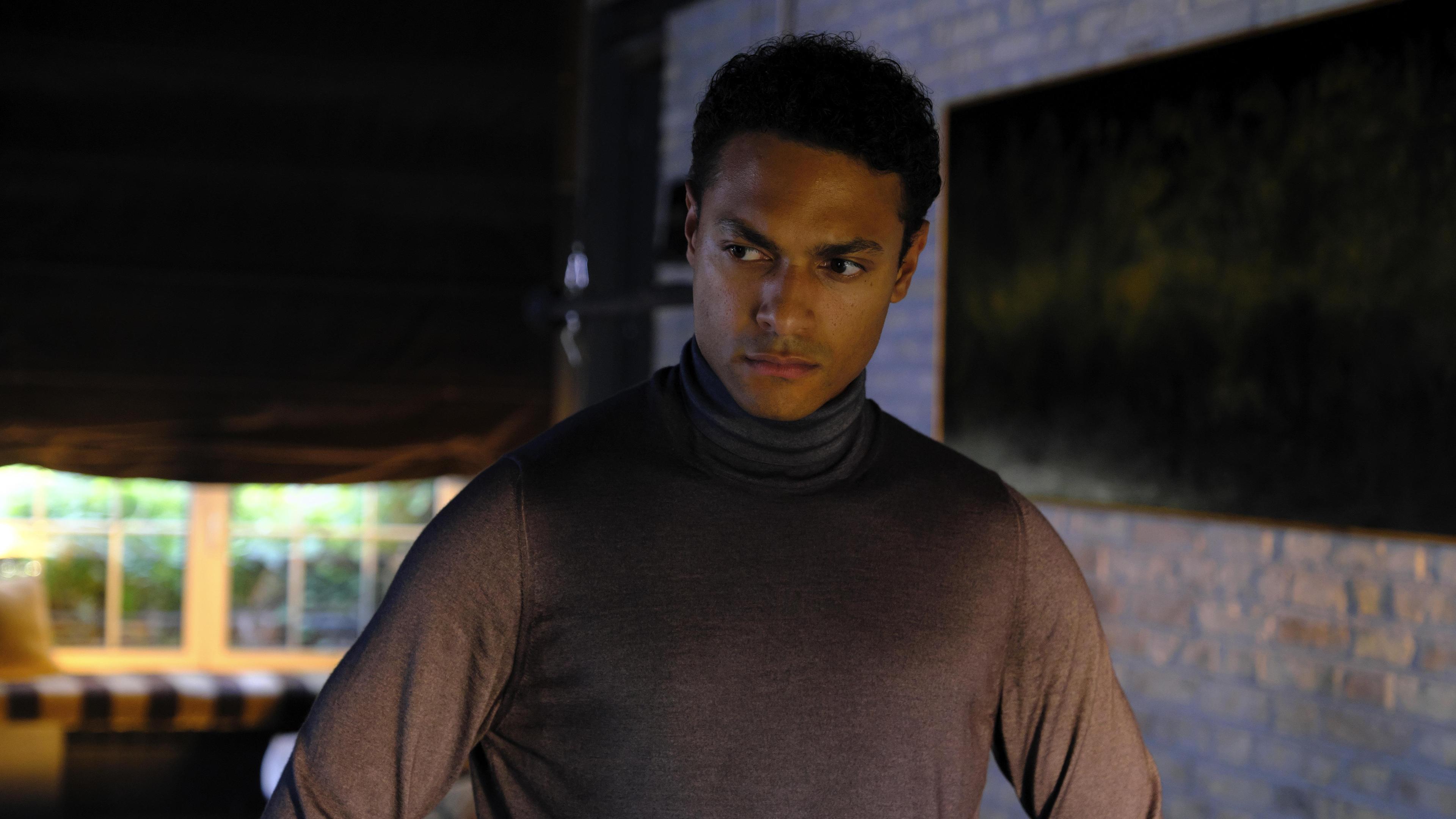 Bild aus der Serie Concordia: Noah Ericksen (Steven Sowah), ein junger Mann in einem braunen Rollkragenpulli hat die Hände in die Hüften gestemmt und sieht nachdenklich aus.