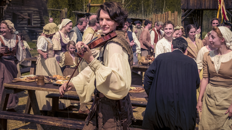 Auf dem Dorfplatz sind viele Reihen von Bänken und Tischen voller Speisen aufgestellt. Viele gut gelaunte Menschen in bäuerlicher Tracht bevölkern das Bild, alte und junge, Männer, Frauen und Kinder. Zentral im Vordergrund Rübezahl (Sabin Tambrea) als Jäger Montanus. Er trägt eine schneeweiße Bluse, Wams und Hose in Leder. Ausgelassen spielt er auf der Geige.