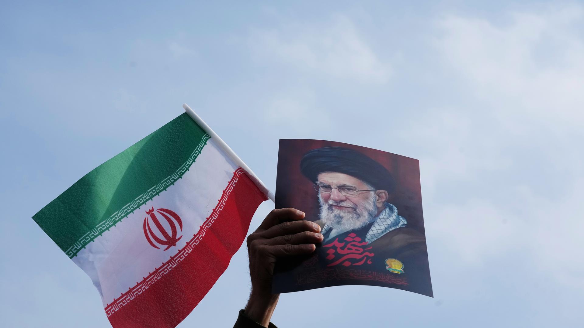 Eine Hand hält eine iranische Flagge und ein Bild vom getöteten iranischen Führer Ajatollah Ali Chamenei hoch.