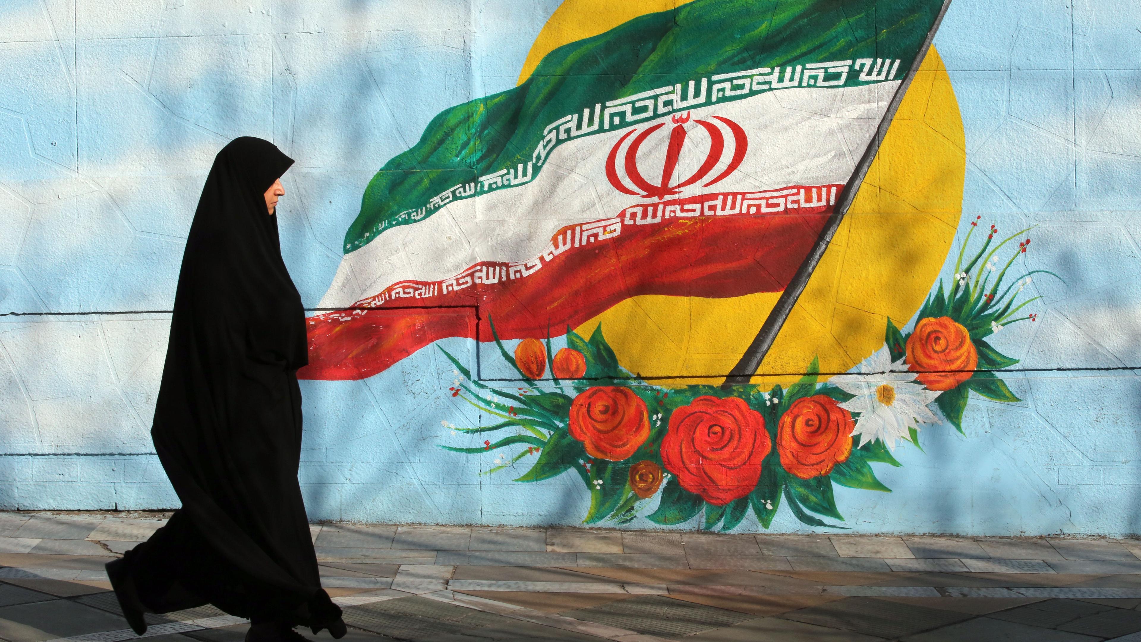 Frau mit Kopftuch läuft vor einer Wand, wo eine iranische Flagge zu sehen ist