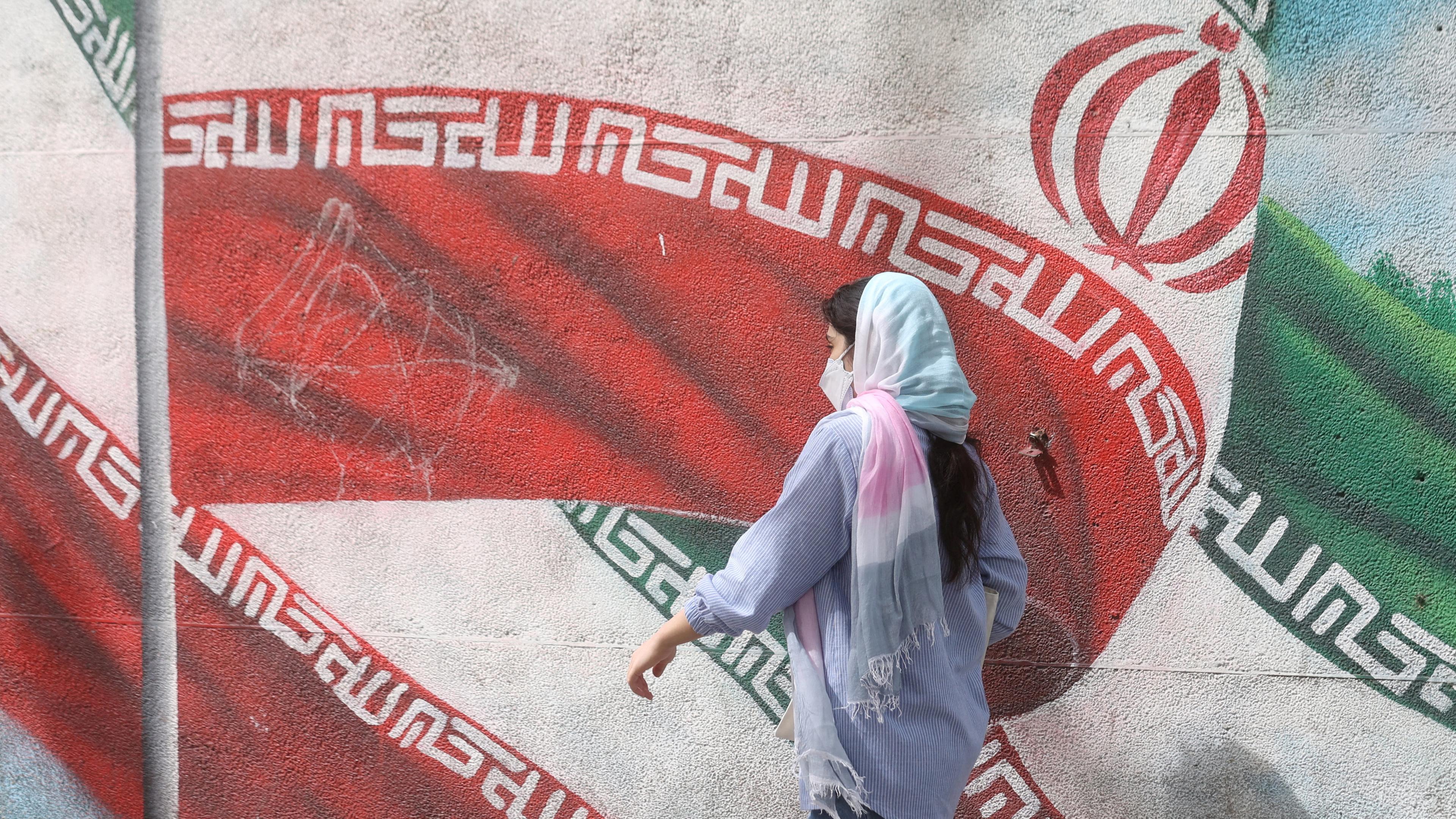 Zu sehen ist eine Frau mit Kopftuch vor einer Mauer in Teheran, auf der die Iran-Flagge aufgemalt ist.