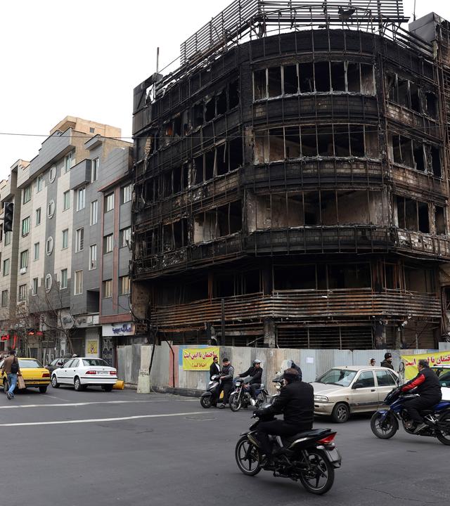 Über eine Straßenkreuzung an der ein ausgebranntes Geschäftshaus steht, fahren Motorradfahrer und Autos.