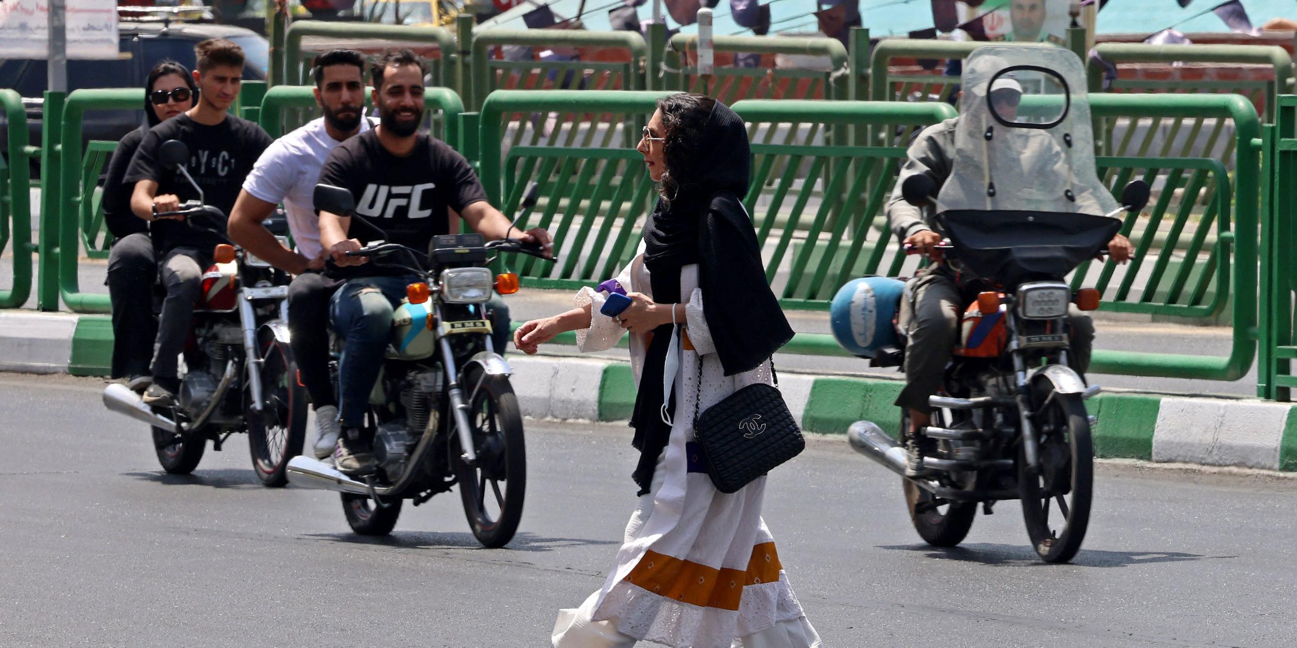 Eine Frau mit Kopftuch überquert die Straße, hinter ihr fahren mehrere Männer auf Motorrädern.