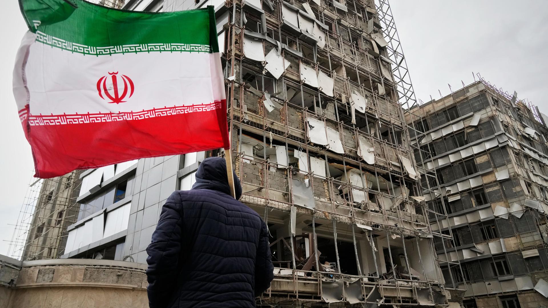 Ein Mann hält eine iranische Fahne neben einem beschädigten Krankenhaus.