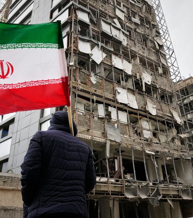 Ein Mann hält eine iranische Fahne neben einem beschädigten Krankenhaus.