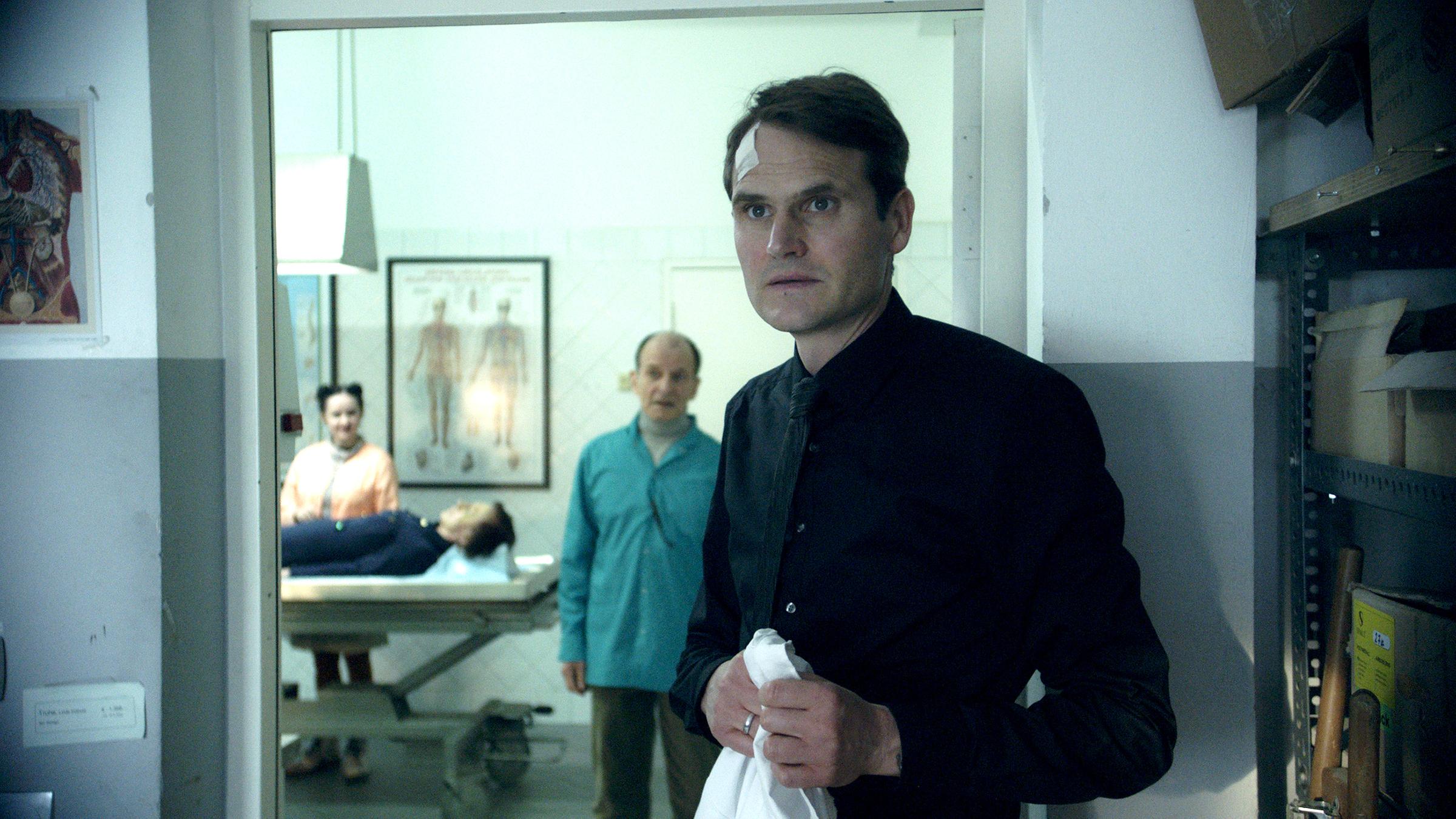 Karsten (Fabian Hinrichs) hat ein weißes Pflaster an der Stirn und hält einen weißen Kittel in Händen. Er steht vor dem Durchgang zu einem Leichenschauraum, in dem aufgebahrt eine Leiche liegt, und starrt ungläubig auf etwas außerhalb des Bildes.