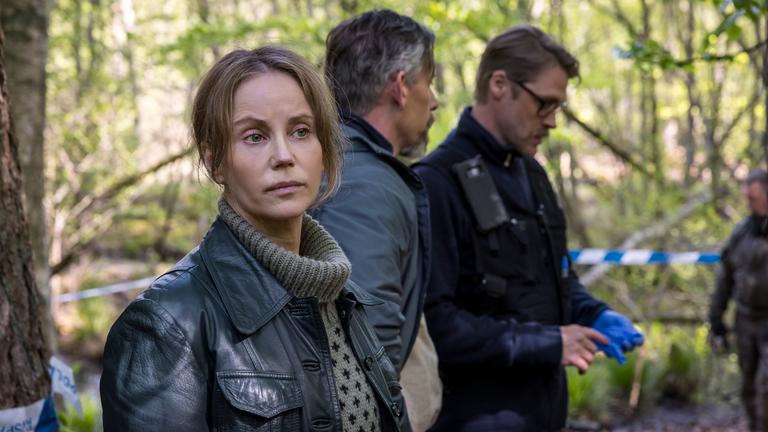 "Iris - Die Wahrheit – Der Fund": Iris (Sofia Helin) schaut sich mit skeptischem Blick im Wald um. Hinter ihr hantiert der Kriminaltechniker Tomas (Magnus Schmitz), der an einer Hand einen Gummihandschuh trägt. Jens (Håkan Bengtsson) sieht Tomas bei der Arbeit zu.