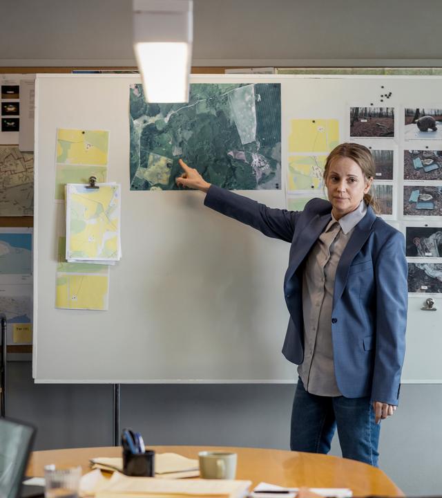 "Iris - Die Wahrheit: Entdeckungen": Iris Broman (Sofia Helin) steht in einem Raum an einer Stellwand. Sie deutet mit dem rechten Zeigefinger auf ein großes Bild, das dort aufgehängt ist. Daneben befinden sich weitere Fotos und Dokumente. Iris gegenüber sitzen zwei Personen an einem Tisch, die nur zum Teil und von hinten zu sehen sind.