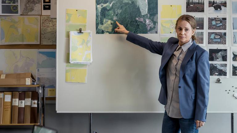 "Iris - Die Wahrheit: Entdeckungen": Iris Broman (Sofia Helin) steht in einem Raum an einer Stellwand. Sie deutet mit dem rechten Zeigefinger auf ein großes Bild, das dort aufgehängt ist. Daneben befinden sich weitere Fotos und Dokumente. Iris gegenüber sitzen zwei Personen an einem Tisch, die nur zum Teil und von hinten zu sehen sind.