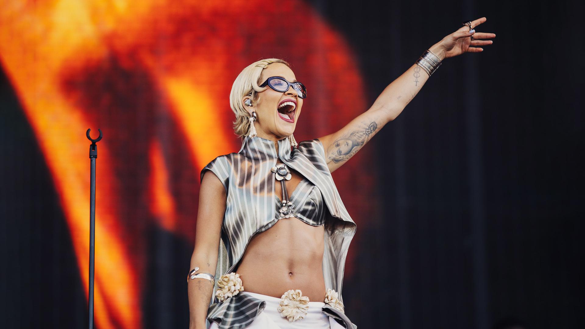 Rita Ora steht lächelnd auf der Bühne und hebt einen Arm in die Luft.