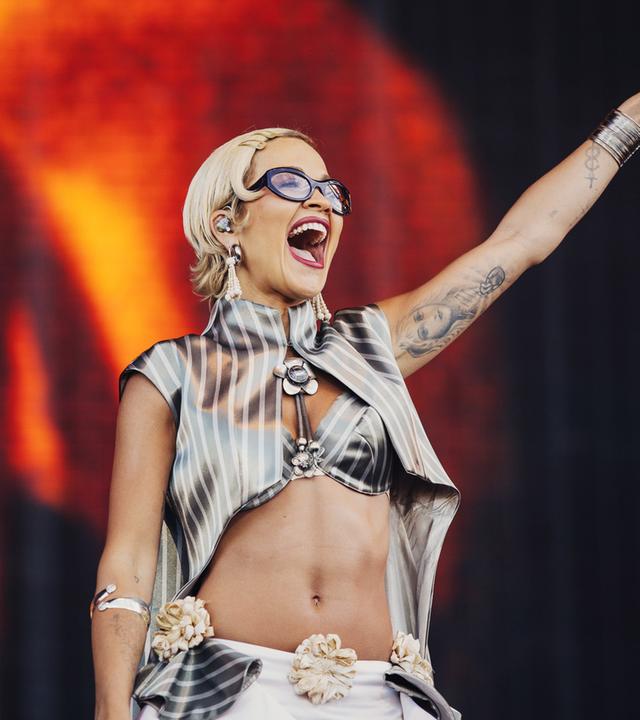 Rita Ora steht lächelnd auf der Bühne und hebt einen Arm in die Luft.