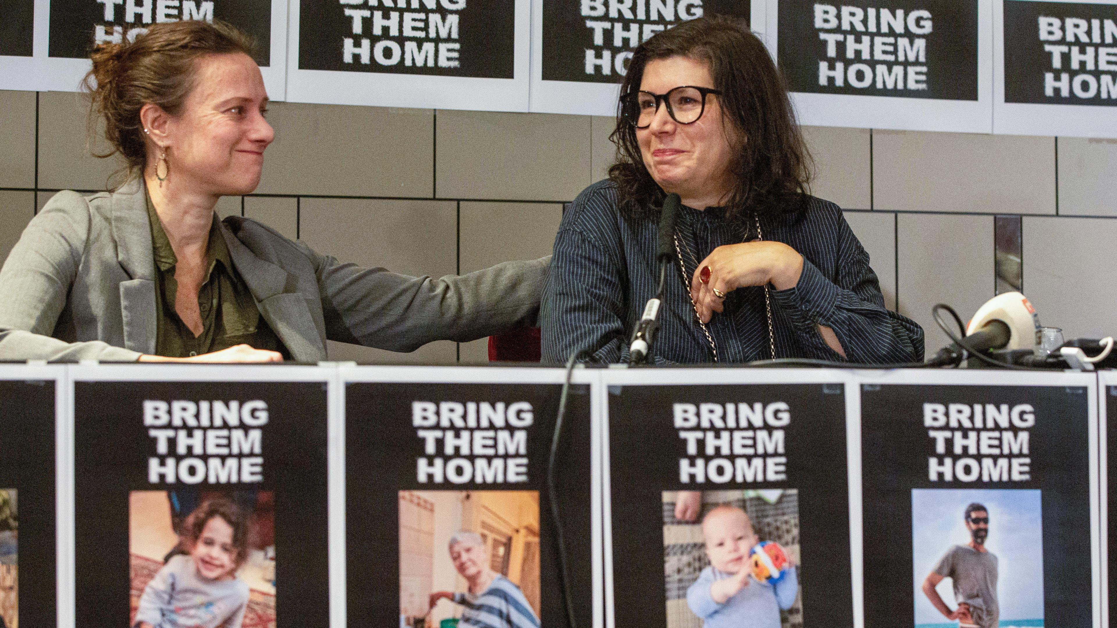Hinter einem Podium, das mit Plakaten von vermissten israelischen Geiseln plakatiert ist, sitzen zwei Frauen. Eine ist den Tränen nahe und wird von einer zweiten getröstet.