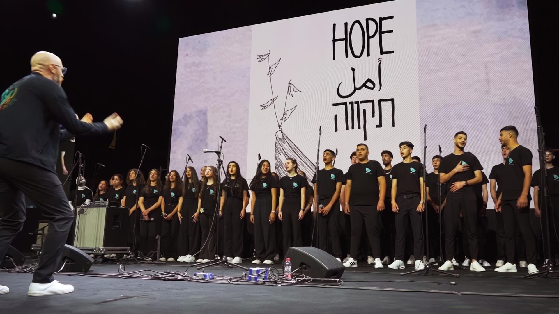 Schwarz gekleidete Jugendliche stehen auf einer Bühne und singen, im Hintergrund wird ein großes weißes Bild mit den Worten Hope eingeblendet.