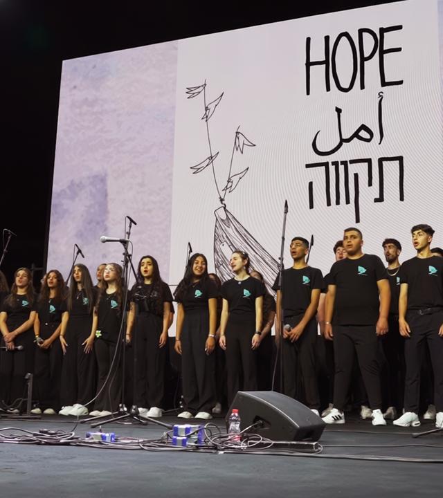 Schwarz gekleidete Jugendliche stehen auf einer Bühne und singen, im Hintergrund wird ein großes weißes Bild mit den Worten Hope eingeblendet.