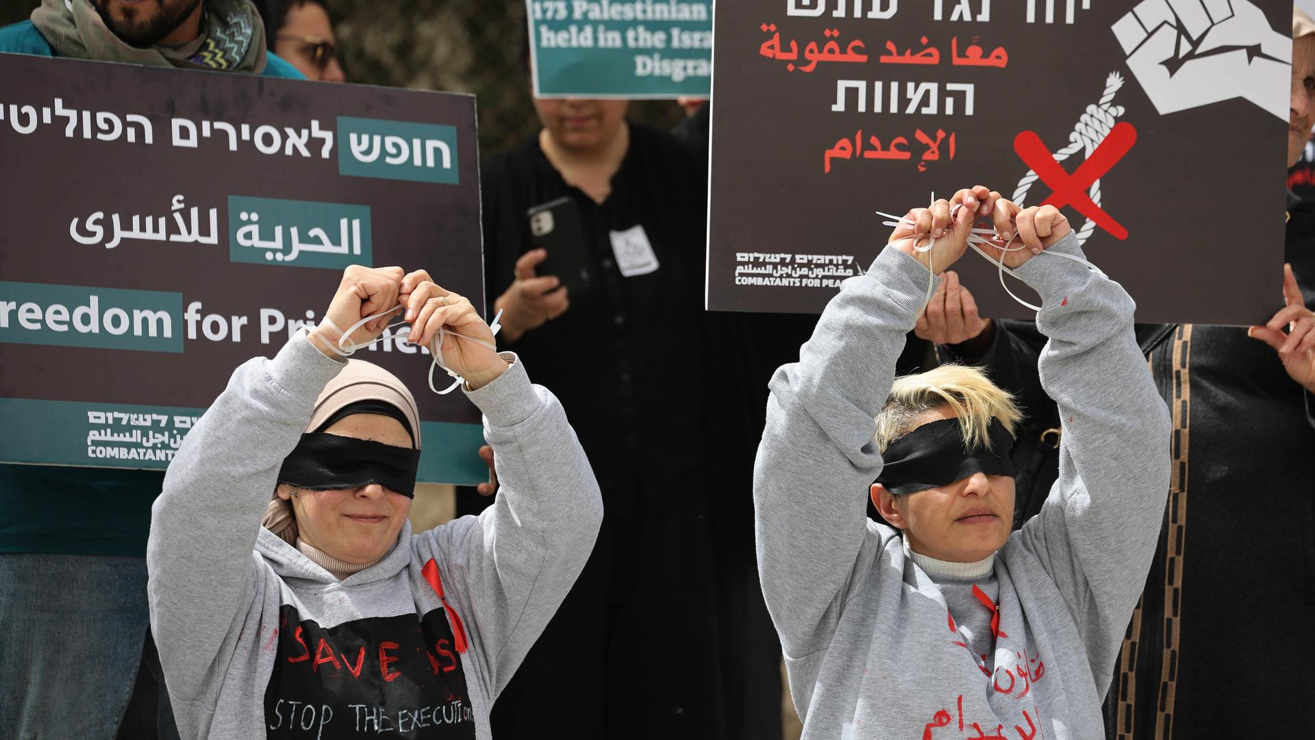 Bei einer Demo halten zwei junge Menschen mit verbundenen Augen ihre gefesselten Hände hoch. Hinter ihnen stehen Menschen mit Plakaten, auf denen etwas in hebräischer Schrift geschrieben steht.
