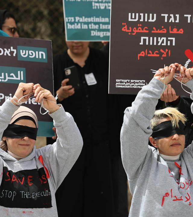 Bei einer Demo halten zwei junge Menschen mit verbundenen Augen ihre gefesselten Hände hoch. Hinter ihnen stehen Menschen mit Plakaten, auf denen etwas in hebräischer Schrift geschrieben steht.