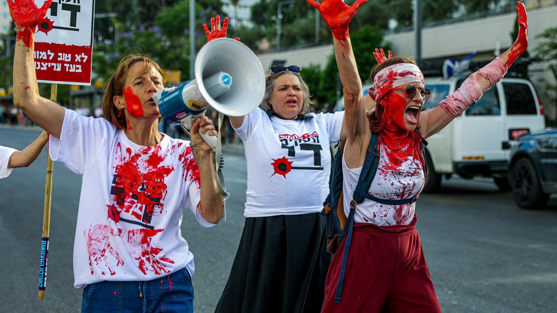Drei Frauen, die sich mit roter Farbe beschmiert haben, laufen schreiend, mit hocherhobenen Händen bei einer Demonstration auf der Straße.