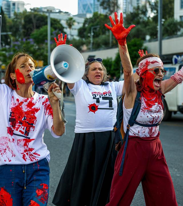 Drei Frauen, die sich mit roter Farbe beschmiert haben, laufen schreiend, mit hocherhobenen Händen bei einer Demonstration auf der Straße.
