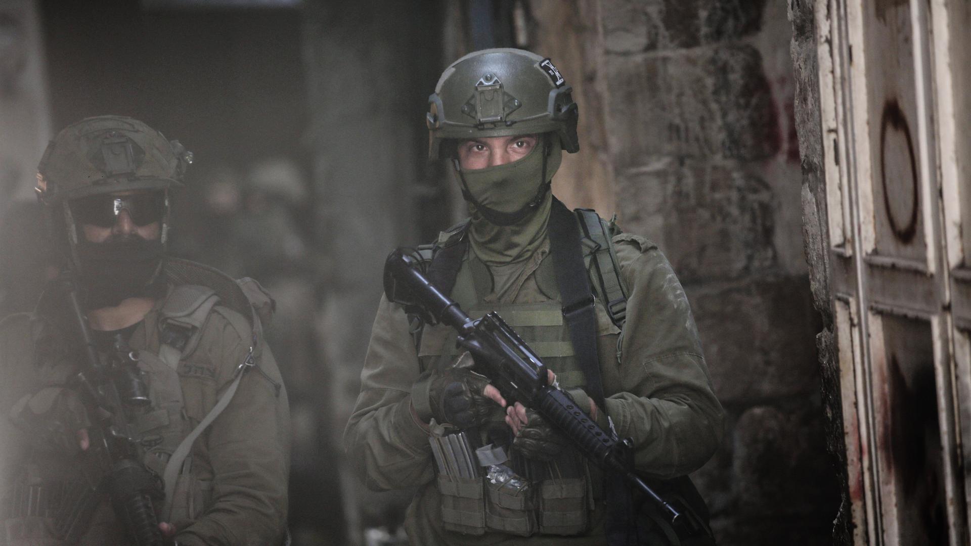 Nahaufnahme zweier Soldaten in grüner Kampfmontur mit vermummten Gesichtern und Gewehren gehen durch eine dunkle Straße.
