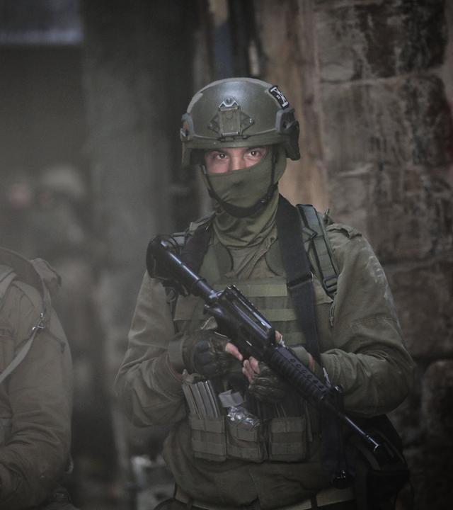 Nahaufnahme zweier Soldaten in grüner Kampfmontur mit vermummten Gesichtern und Gewehren gehen durch eine dunkle Straße.
