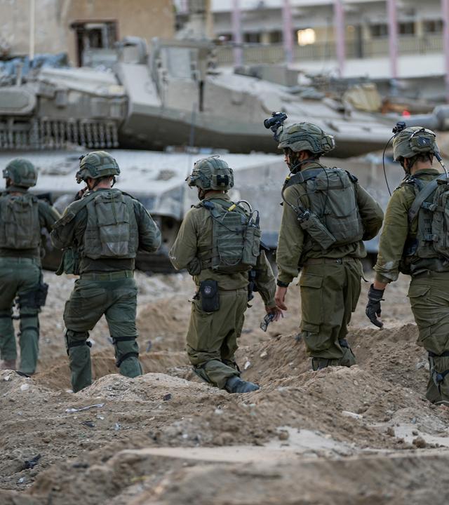 Eine Gruppe Soldaten in armeegrüner Kampfausrüstung läuft durch Schutt. Im Hintergrund sind ein Panzer und zerstörte Gebäude zu sehen