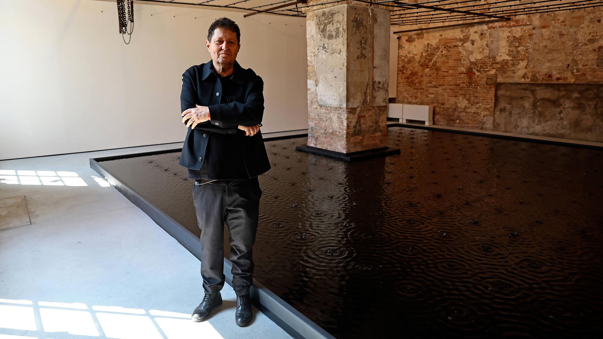 Ein Mann im dunklen Anzug steht mit verschränkten Händen vor einer Wasserinstallation in einem großen Raum mit massiven Steinsäulen.