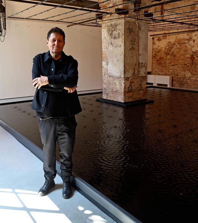 Ein Mann im dunklen Anzug steht mit verschränkten Händen vor einer Wasserinstallation in einem großen Raum mit massiven Steinsäulen.