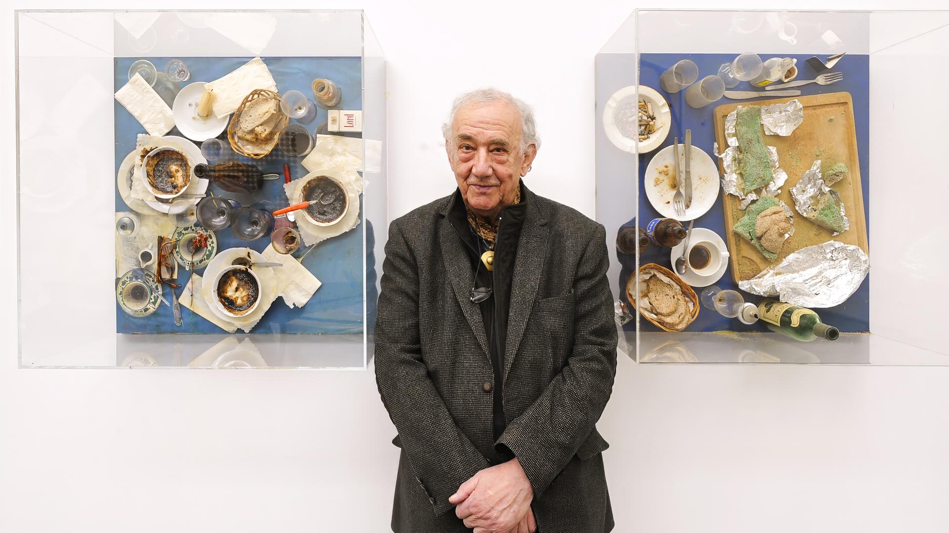 Der Künstler Daniel Spoerri steht zwischen zwei seiner Kunstwerken: der EatArt. Er trägt ein dunklesJackett und ein schwarzes Hemd. Er hält die Hände zusammen vor seinem Bauch. Auf seinen Bildern sind Kaffeetassen, Brot, Aufstriche und Servietten auf einem Tisch zu sehen.