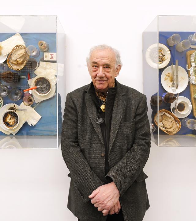 Der Künstler Daniel Spoerri steht zwischen zwei seiner Kunstwerken: der EatArt. Er trägt ein dunklesJackett und ein schwarzes Hemd. Er hält die Hände zusammen vor seinem Bauch. Auf seinen Bildern sind Kaffeetassen, Brot, Aufstriche und Servietten auf einem Tisch zu sehen.