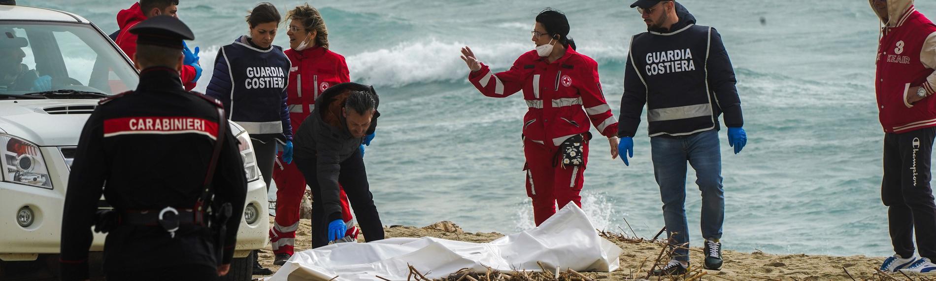 Mitarbeitende des italienischen Roten Kreuzes stehen am Strand und decken eine Leiche ab