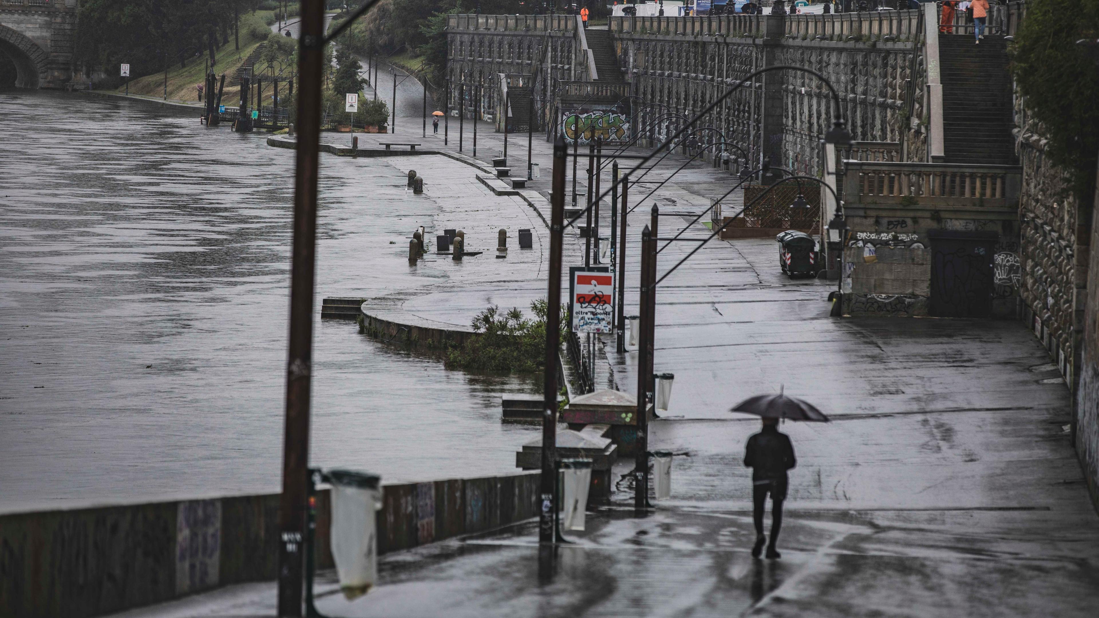 Eine Straße, daneben der Fluss Po. Die Straße steht stark unter Wasser, das Wasser im Fluss steht extrem hoch. Auf der Straße geht ein mann mit einem Regenschirm.